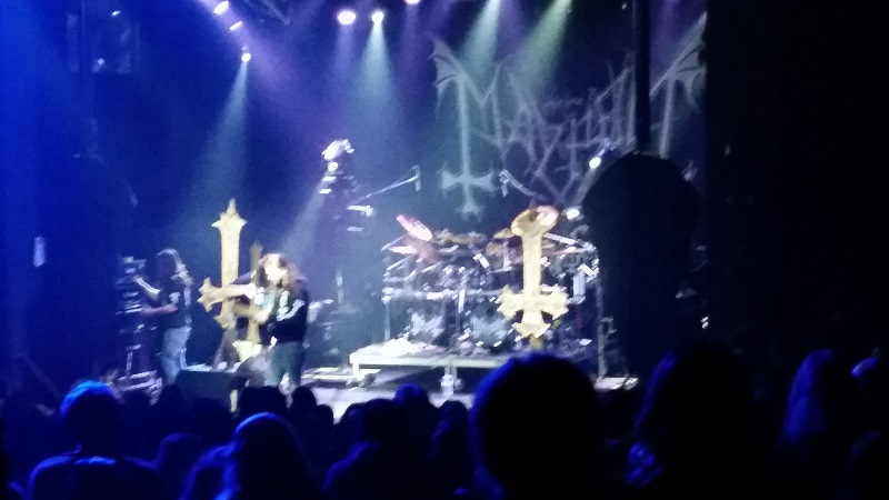 Mayhem's stage, featuring a few upside-down crosses and a monster drum set. A giant banner with Mayhem's band logo hangs in the background. Shafts of stage lighting shine down from above.